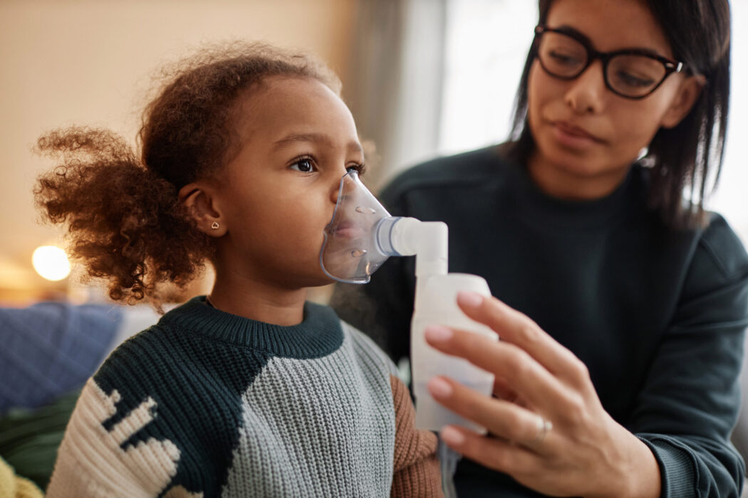 Medium close up shot of little African American girl inhaling medicine through nebulizer while defocused mom holding face mask on daughters face